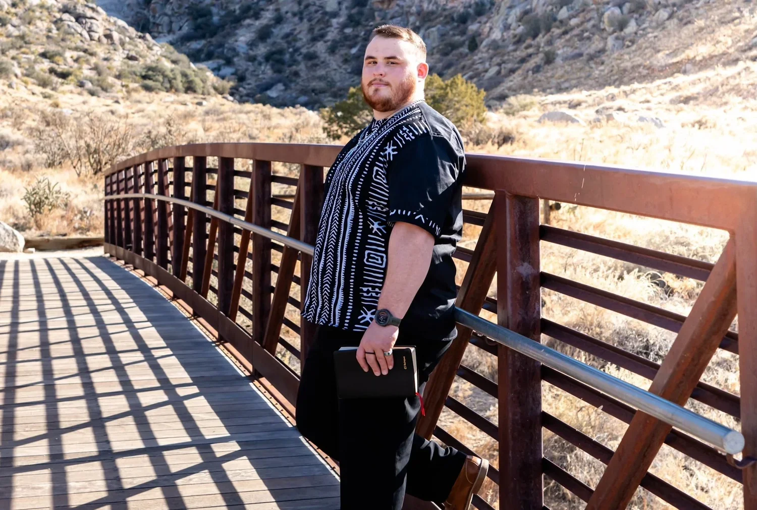 A man with short hair and a beard standing on a bridge in a dry, mountainous landscape. He is wearing a black and white patterned shirt, black pants, and brown shoes. He holds a black notebook or tablet in his left hand and wears a watch on his left wrist. The sun casts striped shadows on the wooden bridge.