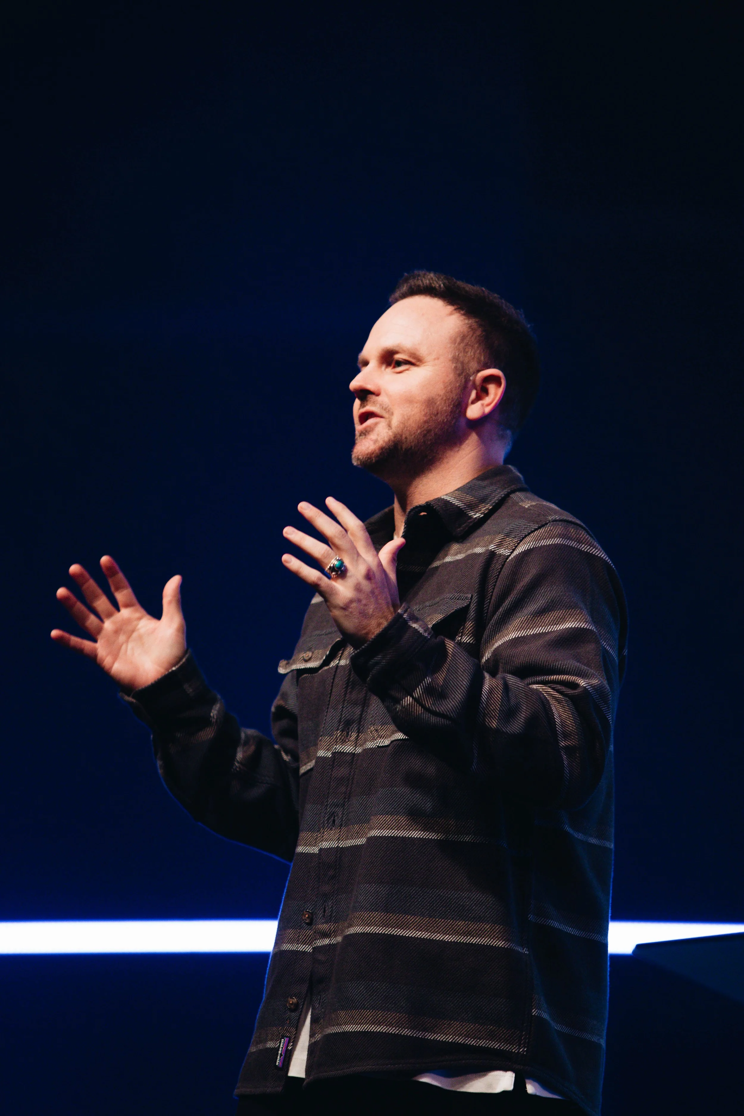 A man with short dark hair and a beard speaking on stage. He is wearing a dark, striped jacket and gesturing with both hands, facing right, against a dark background with blue lighting.