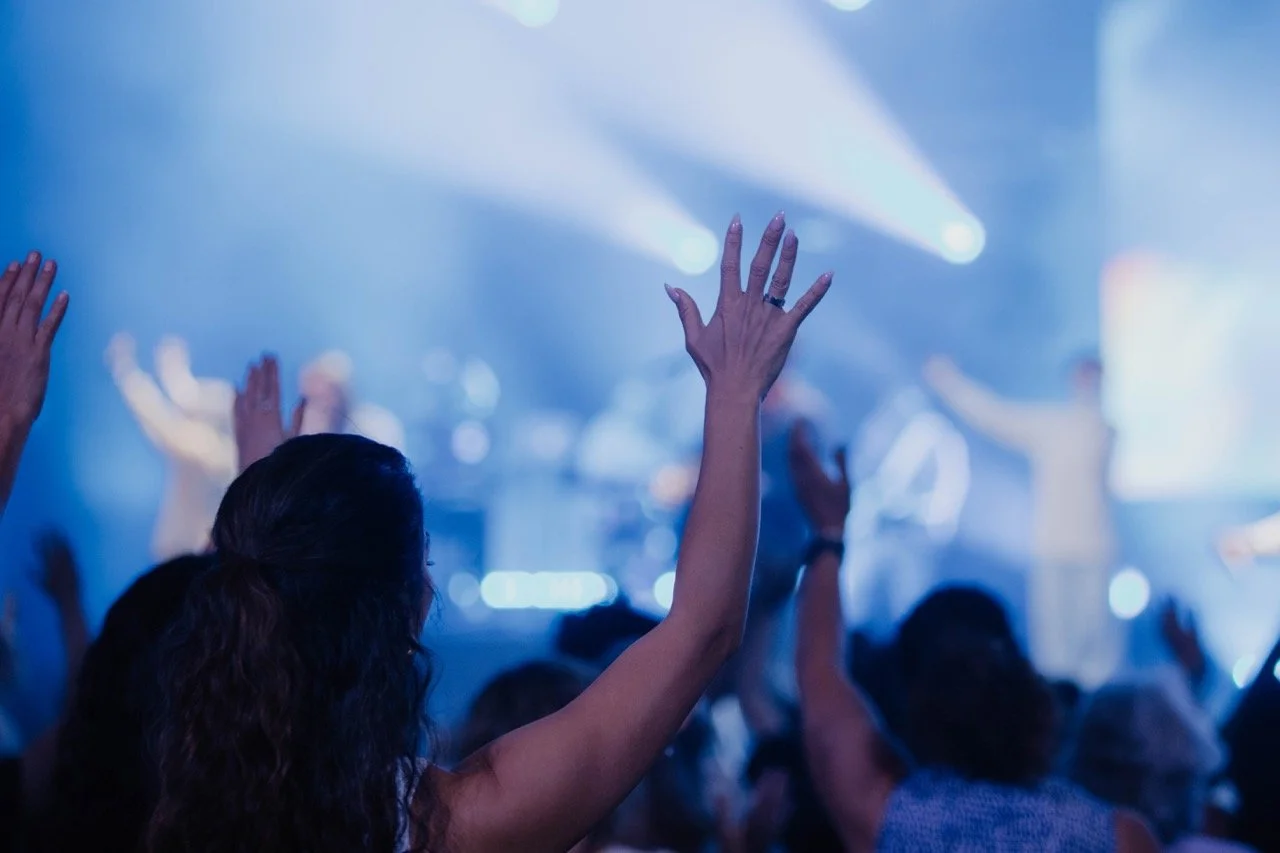 People raising their hands in a concert or worship setting with stage lights and blurred crowd.