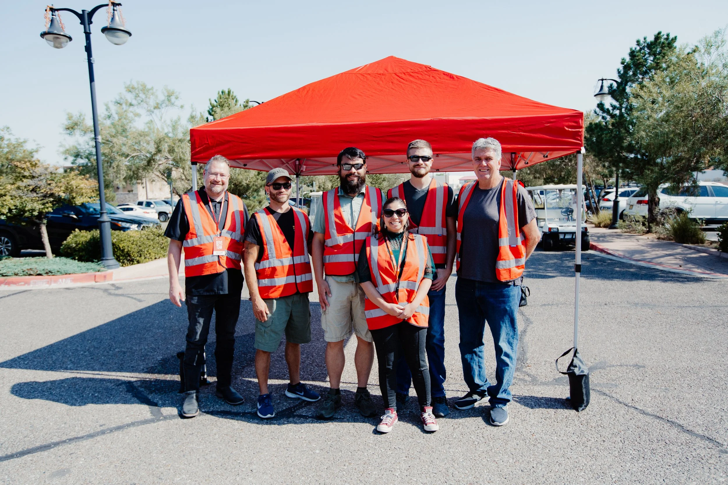Six people standing under a red canopy, wearing orange safety vests, outdoors in a parking lot with trees and cars in the background.