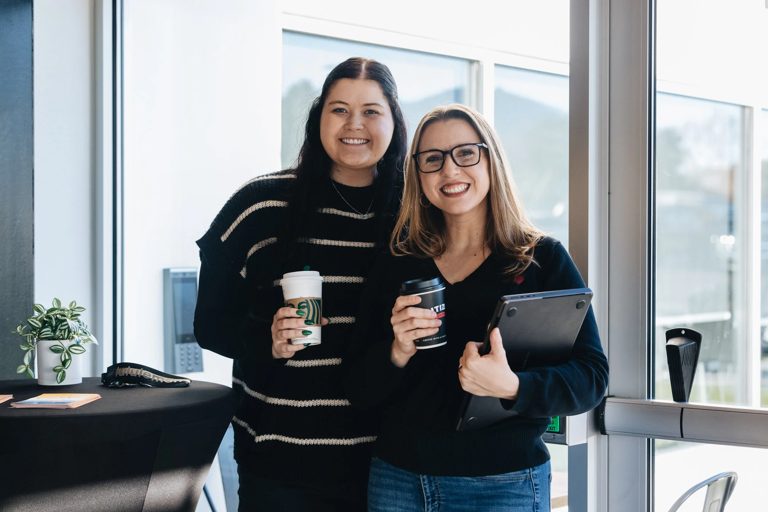 Two women smiling and holding coffee cups, standing near a large window in a modern office space.