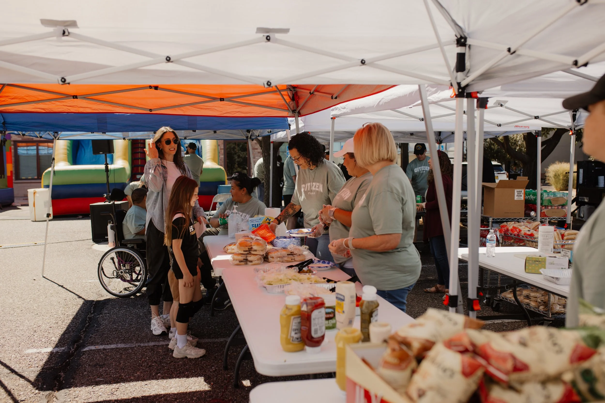 People serving food at an outdoor community event, with tents and inflatable bounce houses in the background.