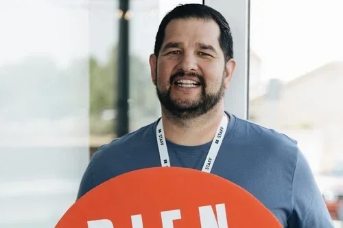 Group of six young adults smiling at the camera indoors, holding cups and snacks, with a mural of a person's face on the wall behind them.