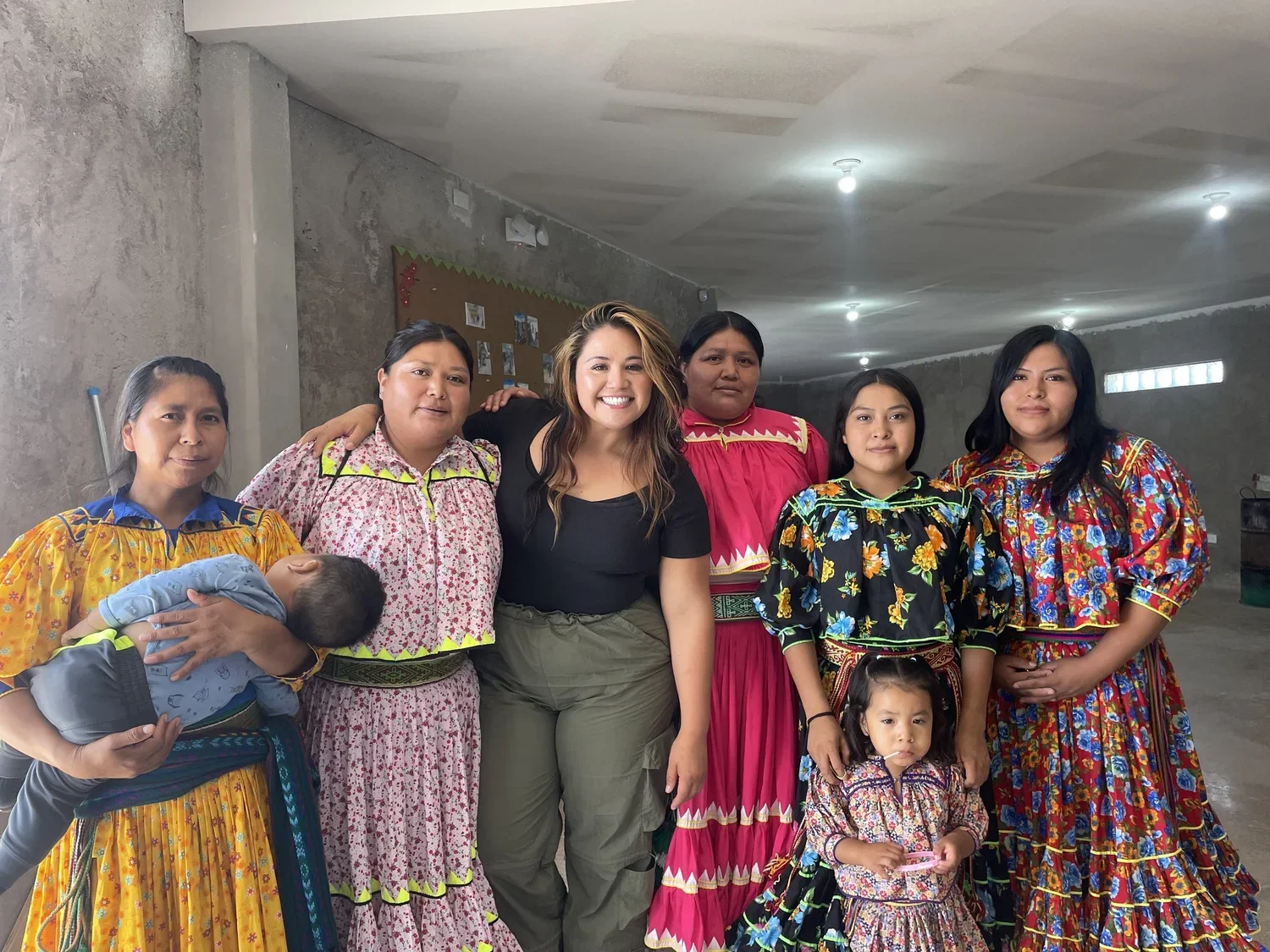 Group of seven women and two children, dressed in colorful traditional Mexican clothing, posing indoors with a plain concrete or plaster wall in the background where there is a bulletin board with photos.