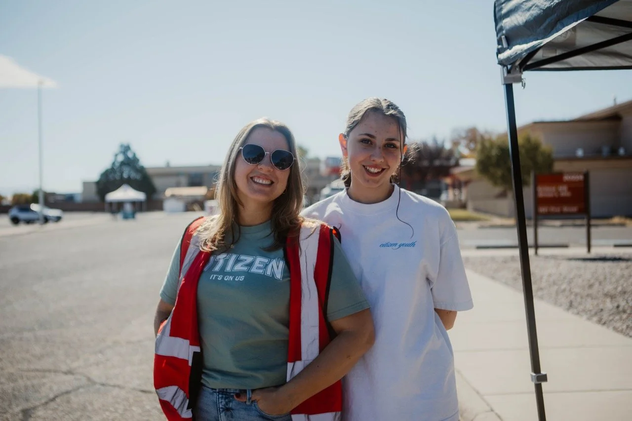 Two young women standing outdoors under a tent, smiling at the camera, with a clear sky and some buildings in the background.
