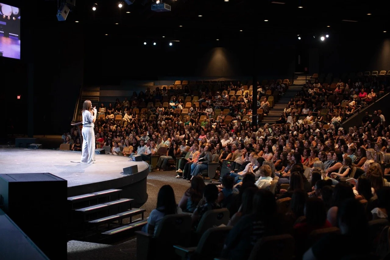 A woman speaking on stage in front of a large audience in a dark auditorium.