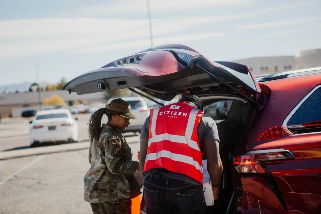 Two people, one in military uniform and one in a red vest with 'Citizen Church' written on the back, are loading supplies into the trunk of a red vehicle in a parking lot.