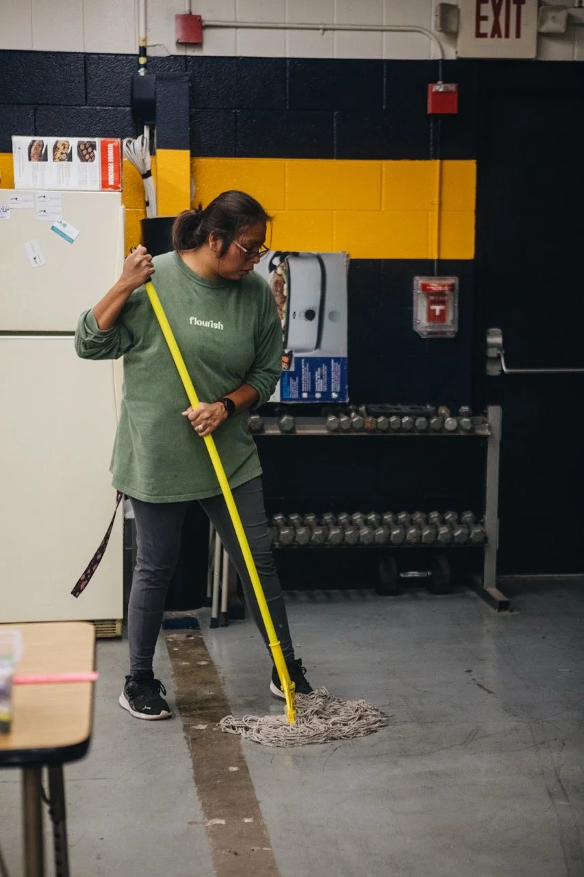 A woman mops the floor in a gym or industrial space, wearing a green sweatshirt and black sneakers, with weights and a poster on the wall behind her.