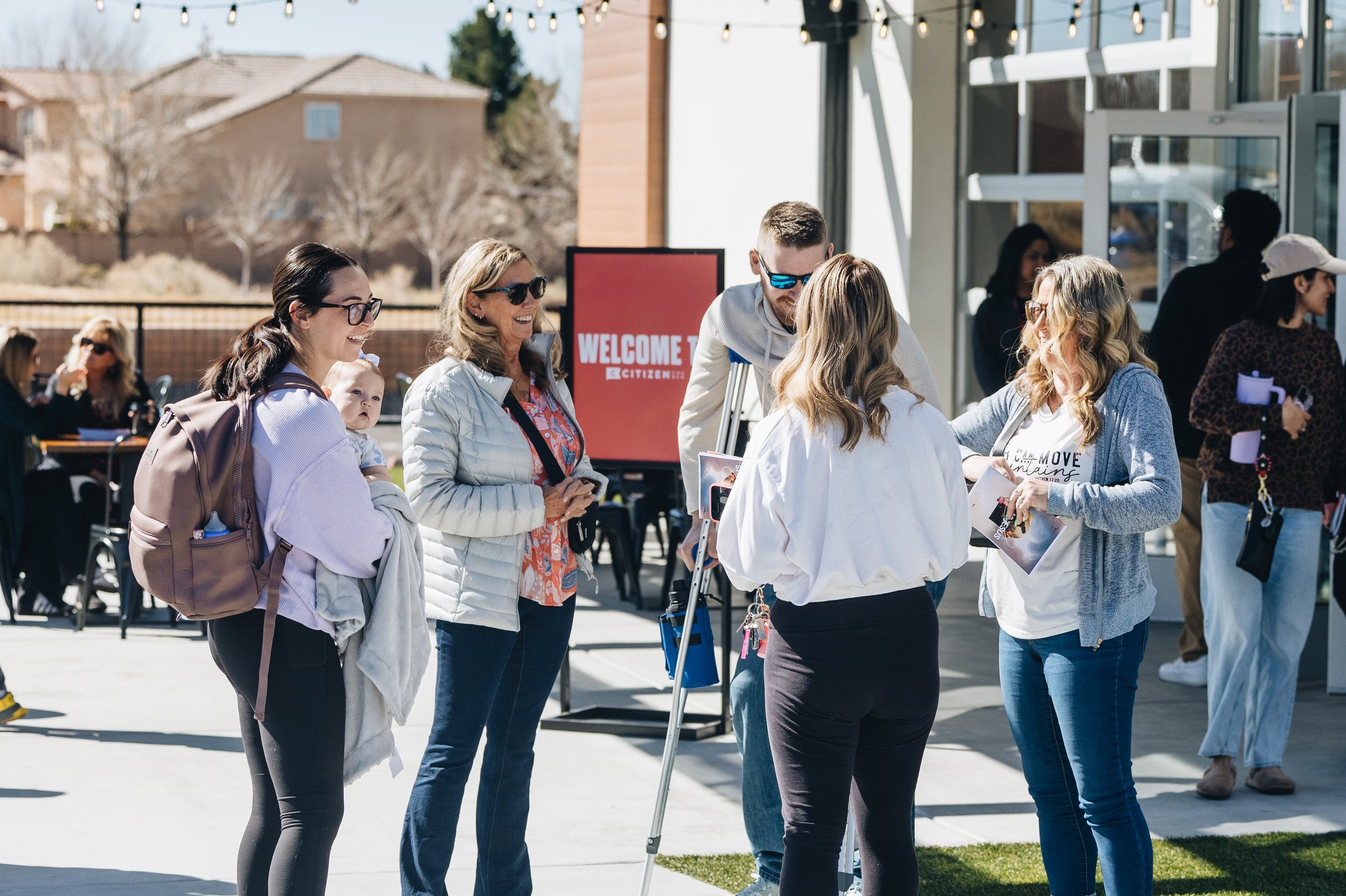 Group of people talking in front of a building with a sign that reads 'Welcome'. Some are smiling and wearing casual clothes, including sunglasses and jackets. A woman is holding a baby, and there are other people in the background. The setting is outside on a sunny day.