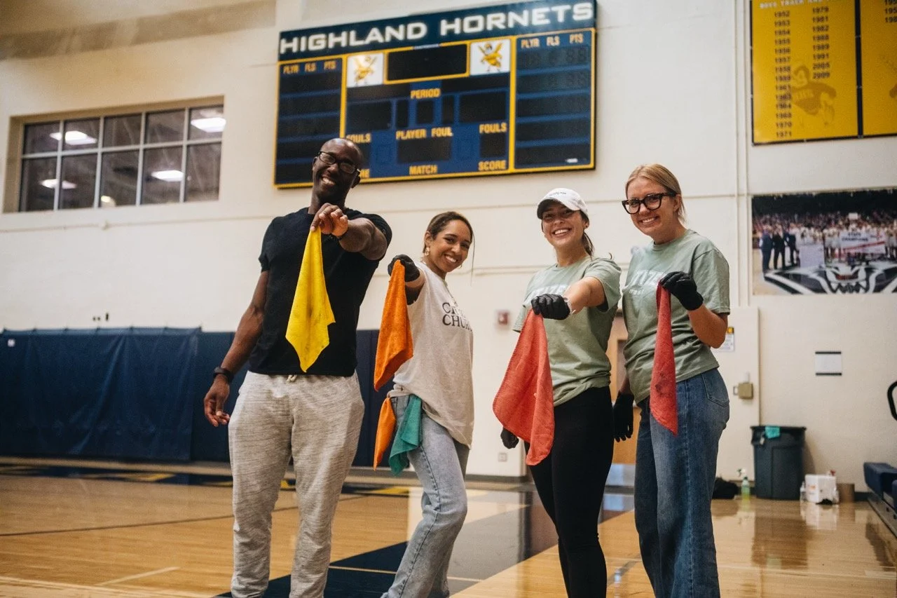 Four people standing in a gymnasium holding orange and yellow towels, smiling and pointing at the camera with a scoreboard in the background reading "Highland Hornets."