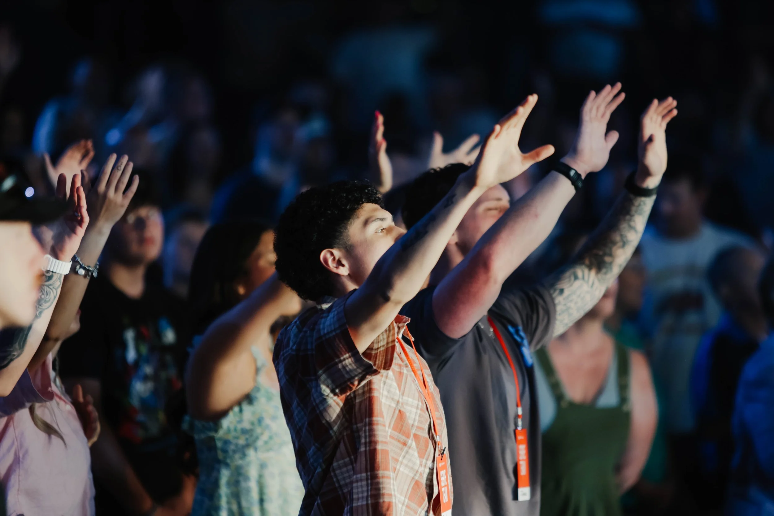 People raising their hands during an event or concert, with some attendees wearing badges around their necks.