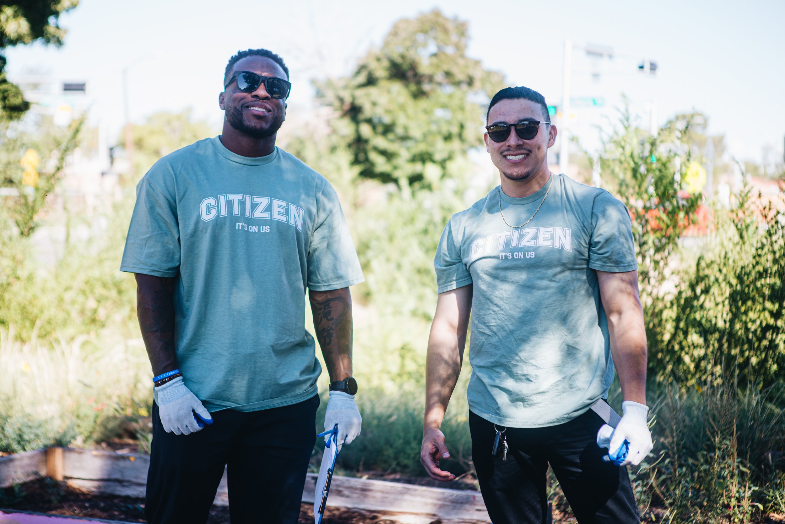 Two men wearing sunglasses and matching green T-shirts that say 'CITIZEN, IT'S ON US' stand outdoors in a garden or park, smiling at the camera.