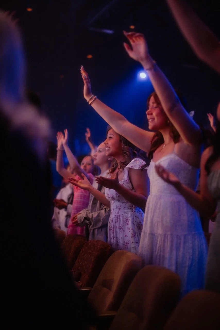 People attending a concert, singing and raising their hands, with stage lighting in the background.