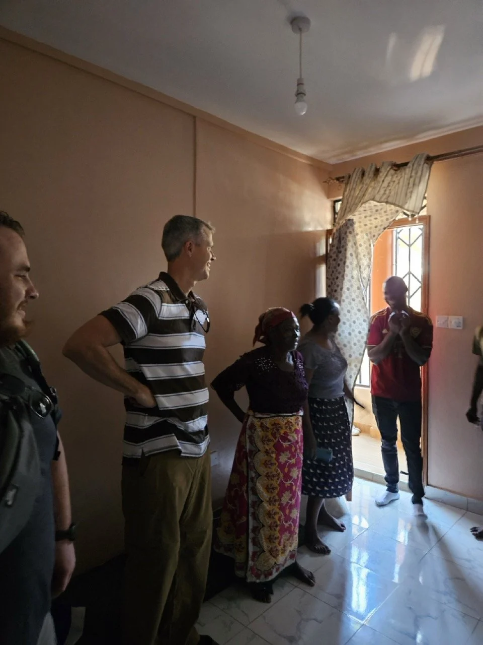 Group of people standing inside a room near an open door with curtains, sunlight coming in, including two women in traditional skirts and a man in a red shirt.