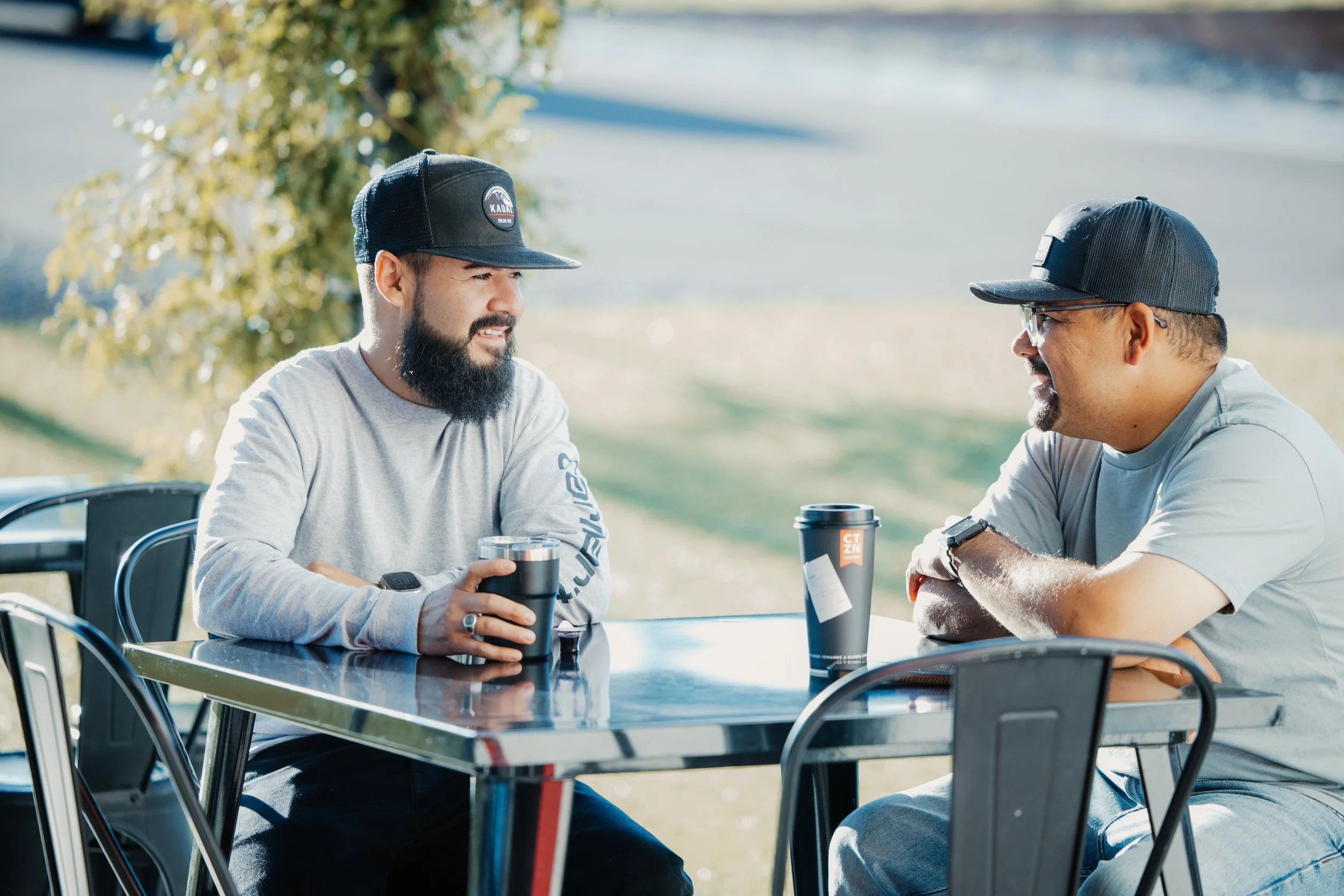 Two men sitting at an outdoor table, conversing and smiling, each with a drink in hand, wearing casual gray T-shirts and black caps, in a park-like setting with green grass and trees in the background.