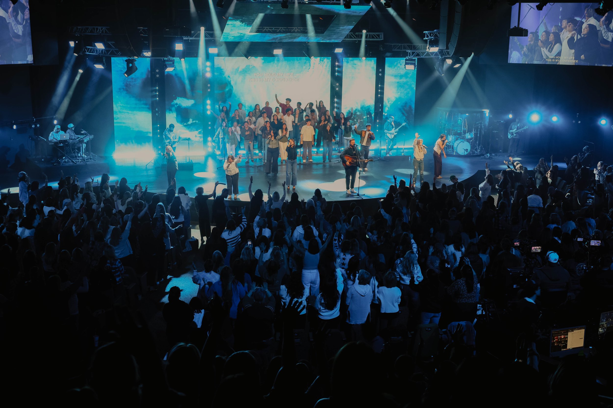 People raising their hands in a concert or worship setting with stage lights and blurred crowd.