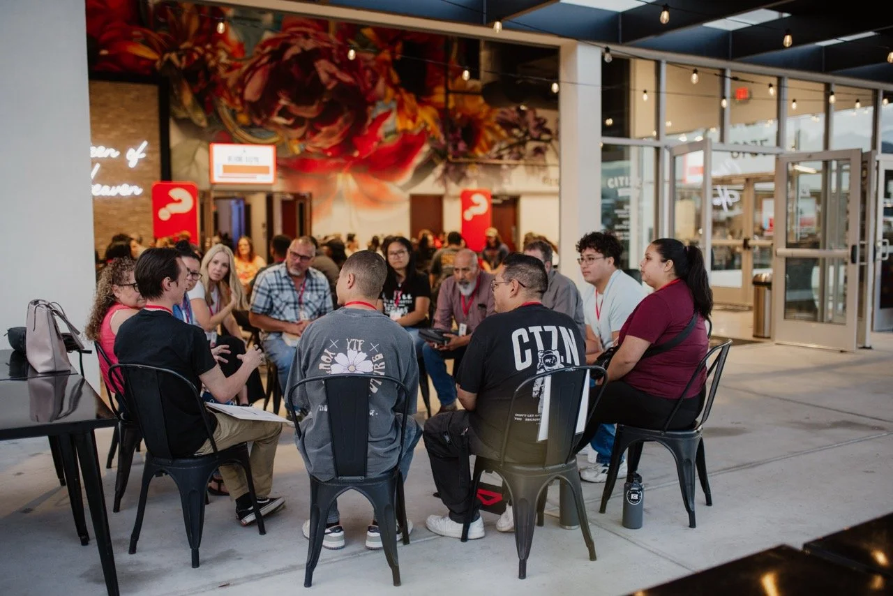 A group of people sitting in a circle at an outdoor event or workshop near a cafe or restaurant.
