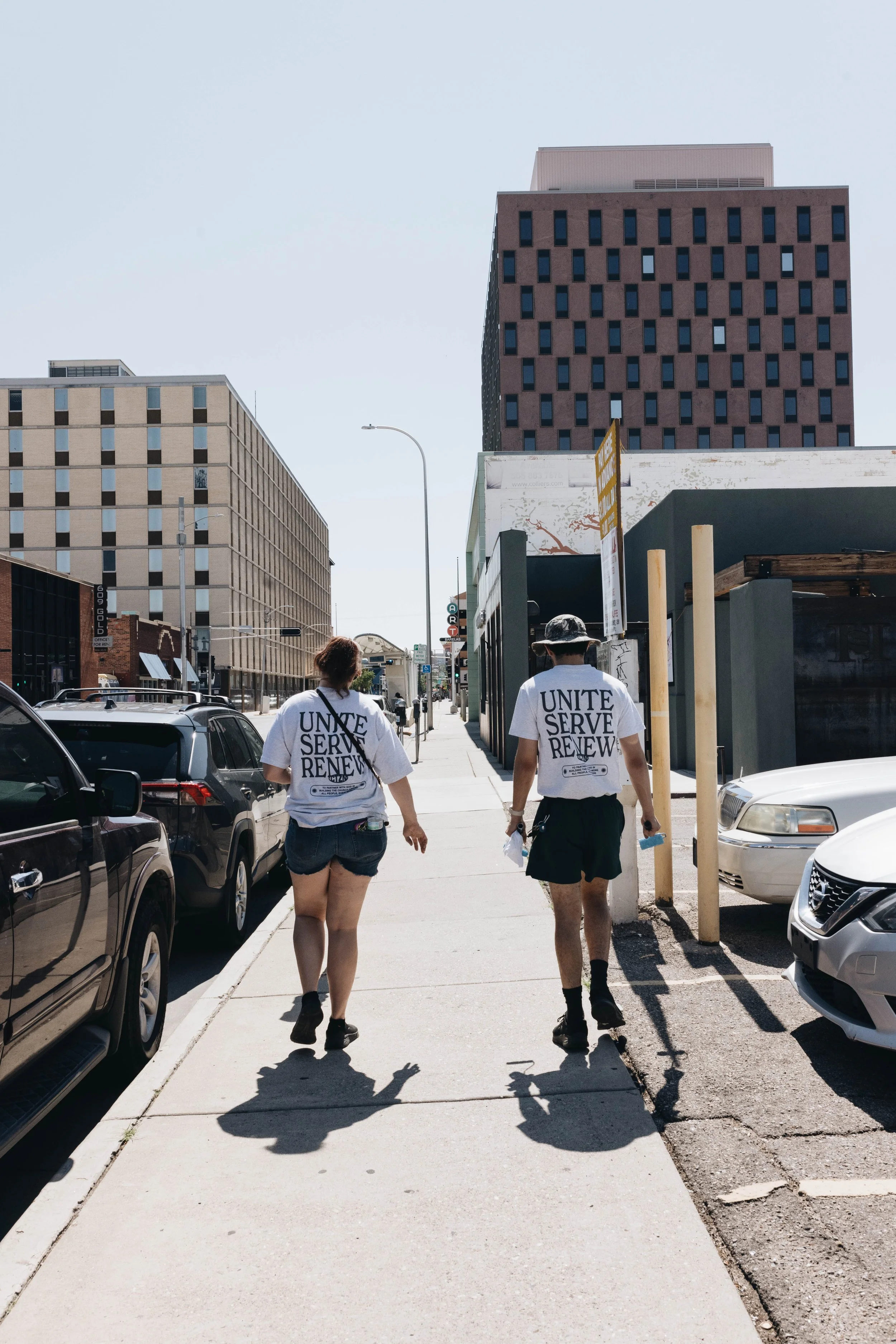 Two people walking on a city sidewalk wearing t-shirts with the words 'UNITE SERVE RENEW'; parked cars line the street, and tall buildings are in the background under a clear sky.