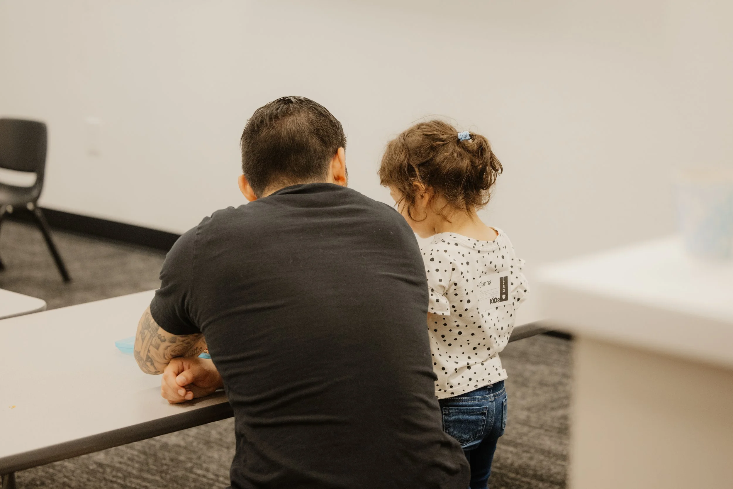A person with short dark hair and a black t-shirt sitting at a table with a young girl with curly brown hair in a white polka dot shirt, in a room with a white wall and some black chairs.