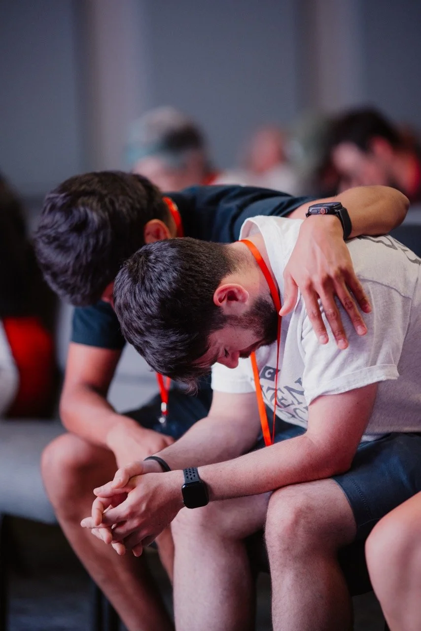 Two young men sitting next to each other, one comforting the other with his arm around his shoulder, in an indoor setting with a blurred background of other people.