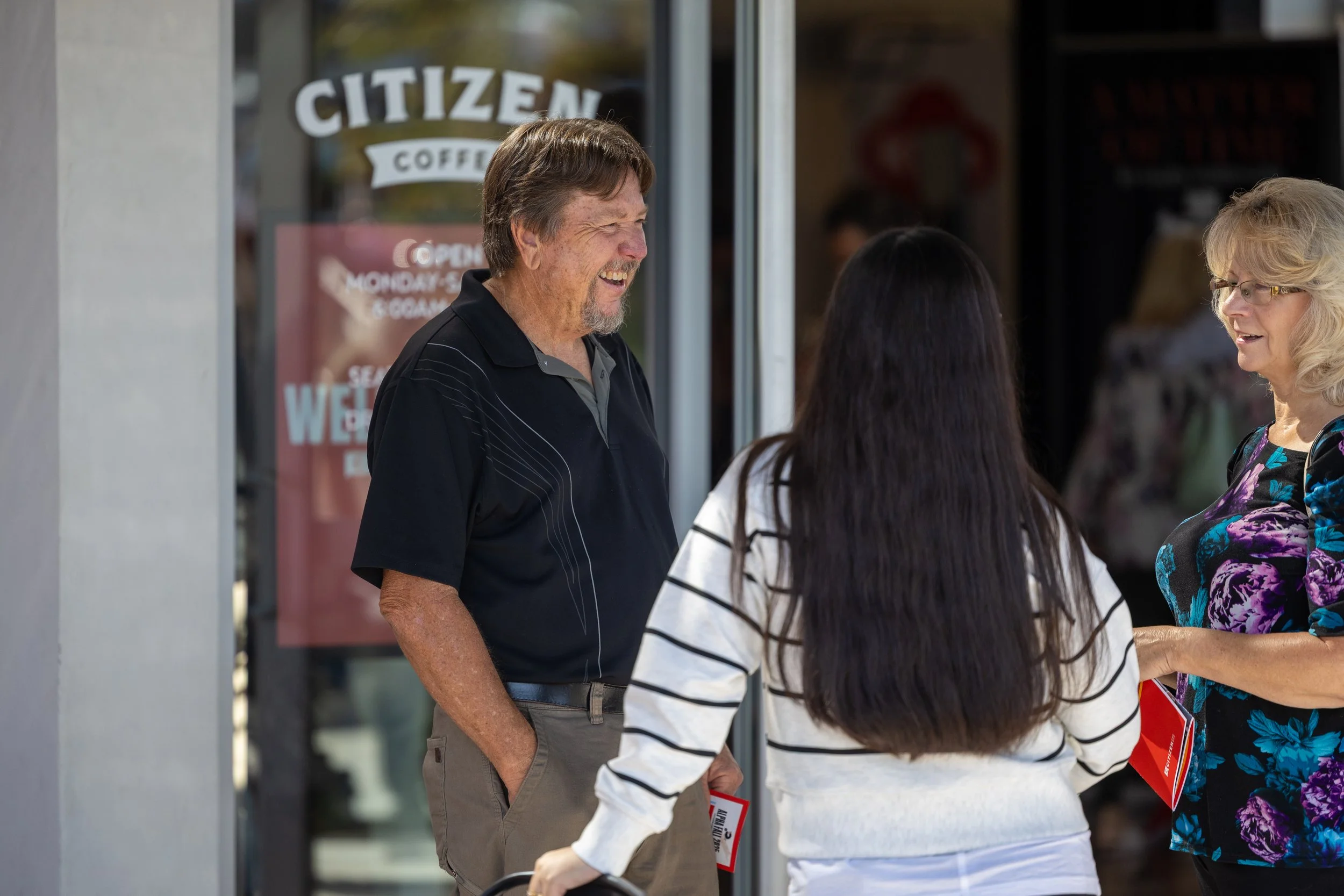 Three people, two women and a man, are having a conversation outside a coffee shop named Citizen Coffee. The man is smiling and wearing a black polo shirt, the woman with long dark hair wears a white shirt with black stripes, and the woman with blonde hair wears glasses and a colorful floral top.