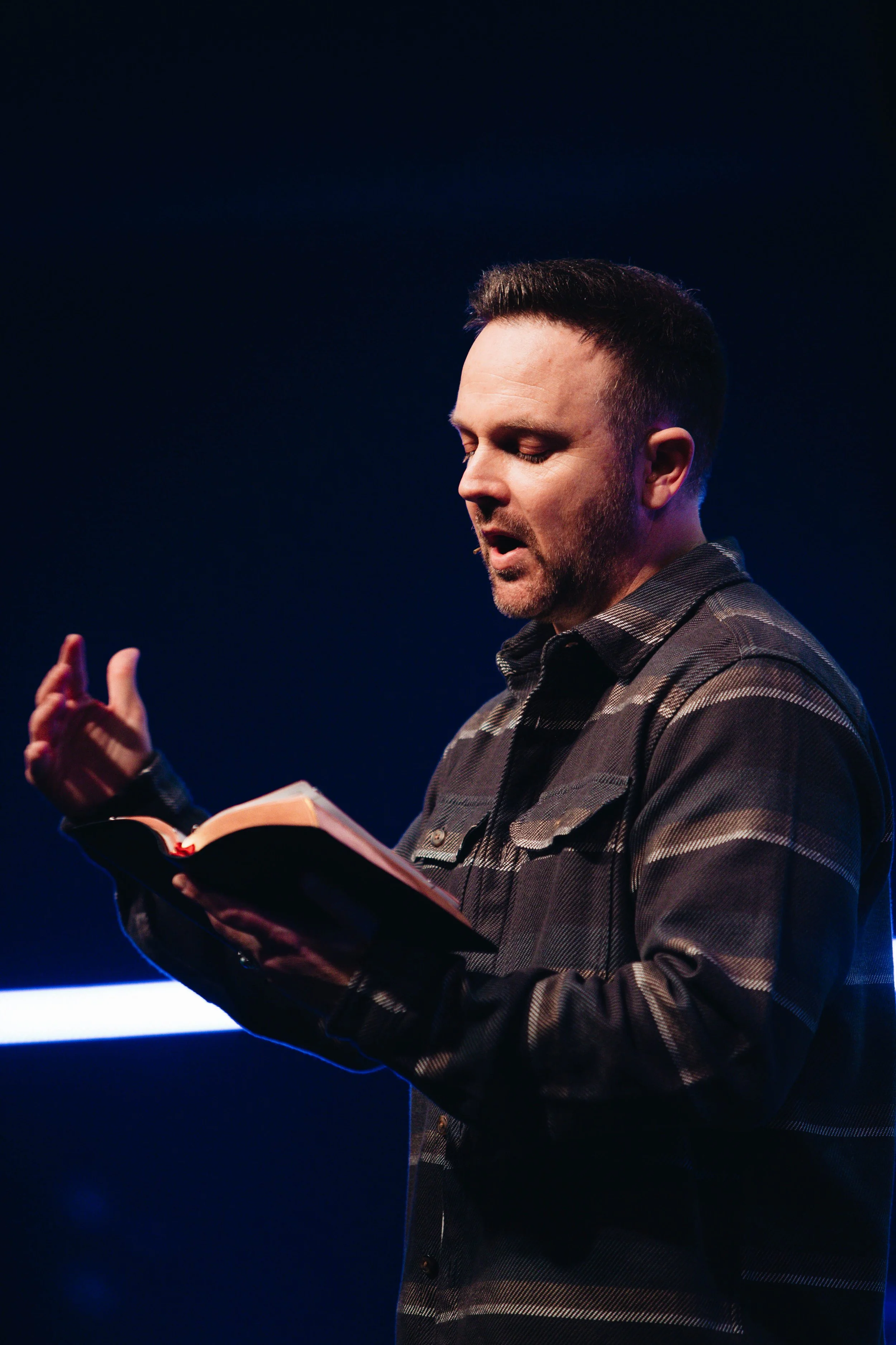 Man with short dark hair and beard reading from a book on stage.