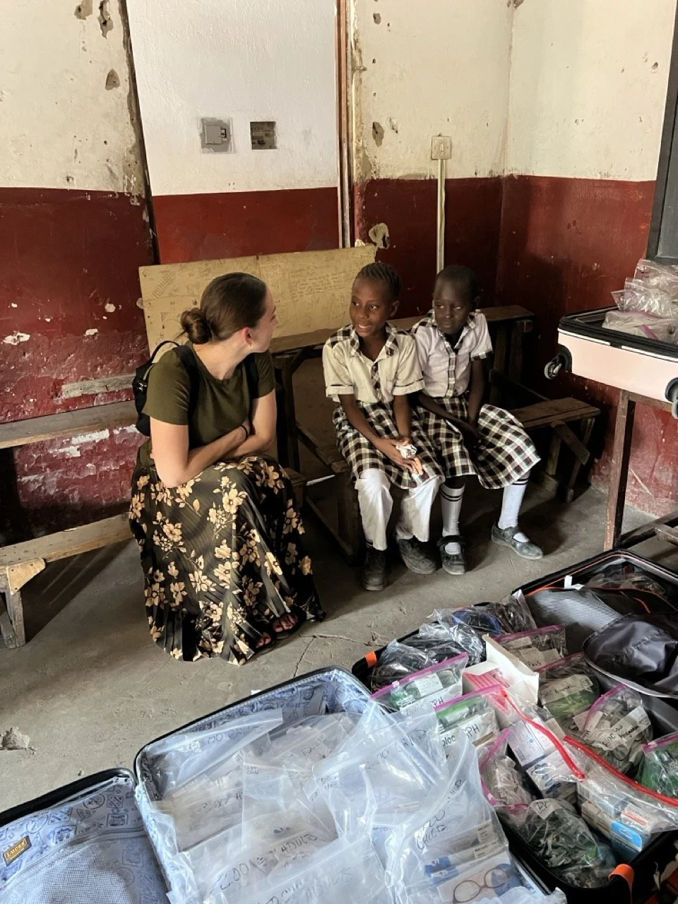 A woman sitting and talking with two young girls in school uniforms inside a classroom with a worn wall and a bench, with various supplies and bags in the foreground.