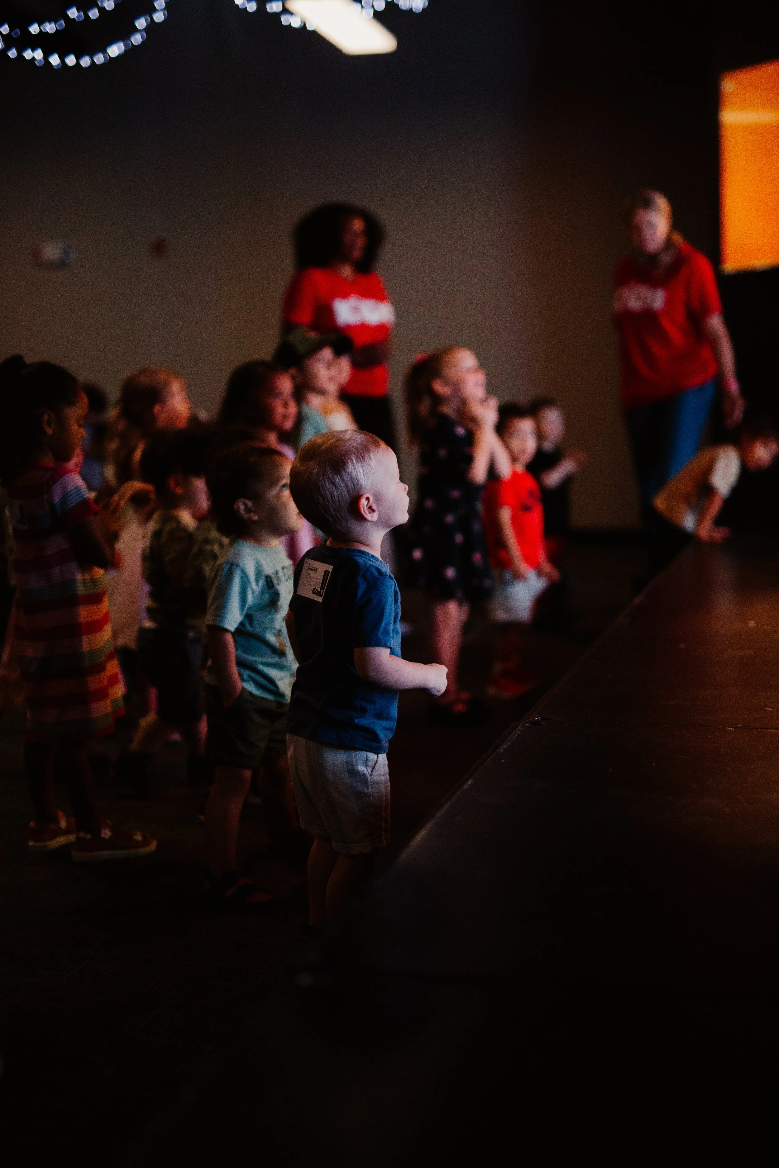 A group of children watching a stage performance in a dark room, with their attention directed towards the stage, lit by minimal lighting.