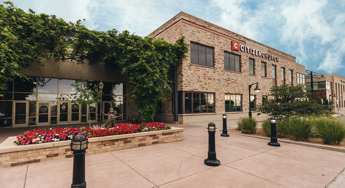Exterior of a brick church building with a sign that reads 'Citizen Church' and a flower bed with pink and white flowers in front.