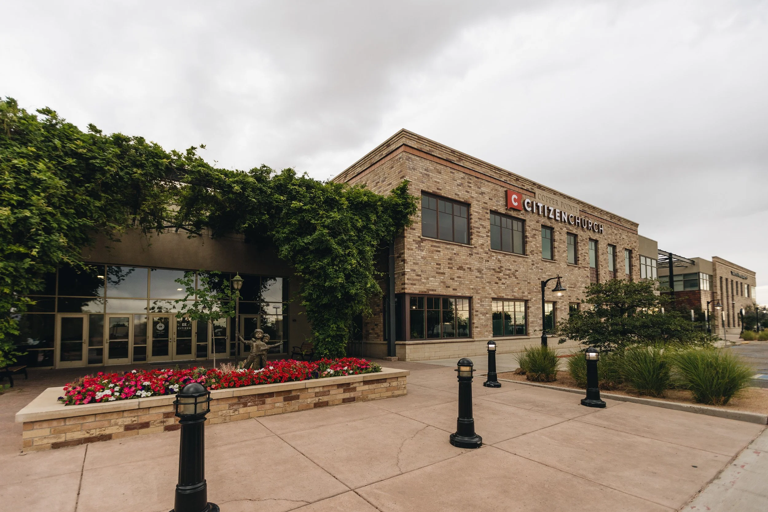Exterior of a modern brick church building named Citizen Church, with a landscaped sidewalk featuring flower beds, small trees, and black bollard lights, under an overcast sky.
