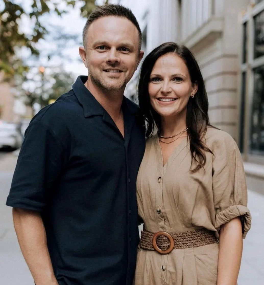 A smiling man and woman standing close together outdoors in an urban setting with trees and buildings in the background.