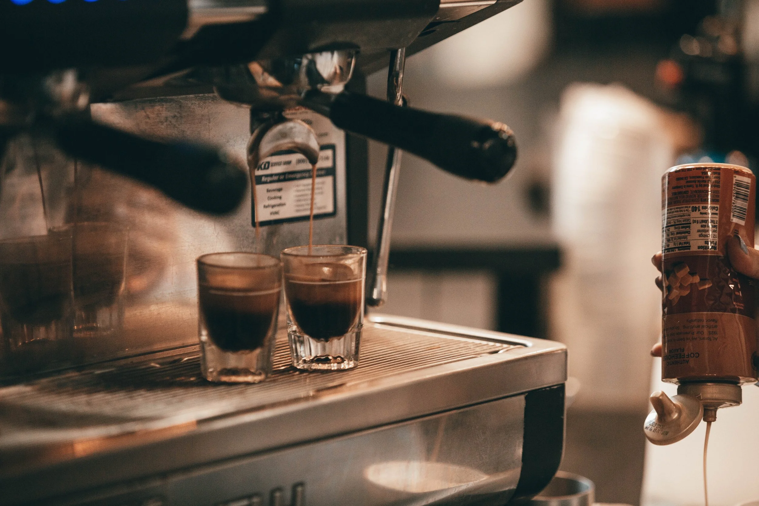 Espresso being poured from a coffee machine into two glass cups, with a person's hand holding a can of coffee creamer on the right.