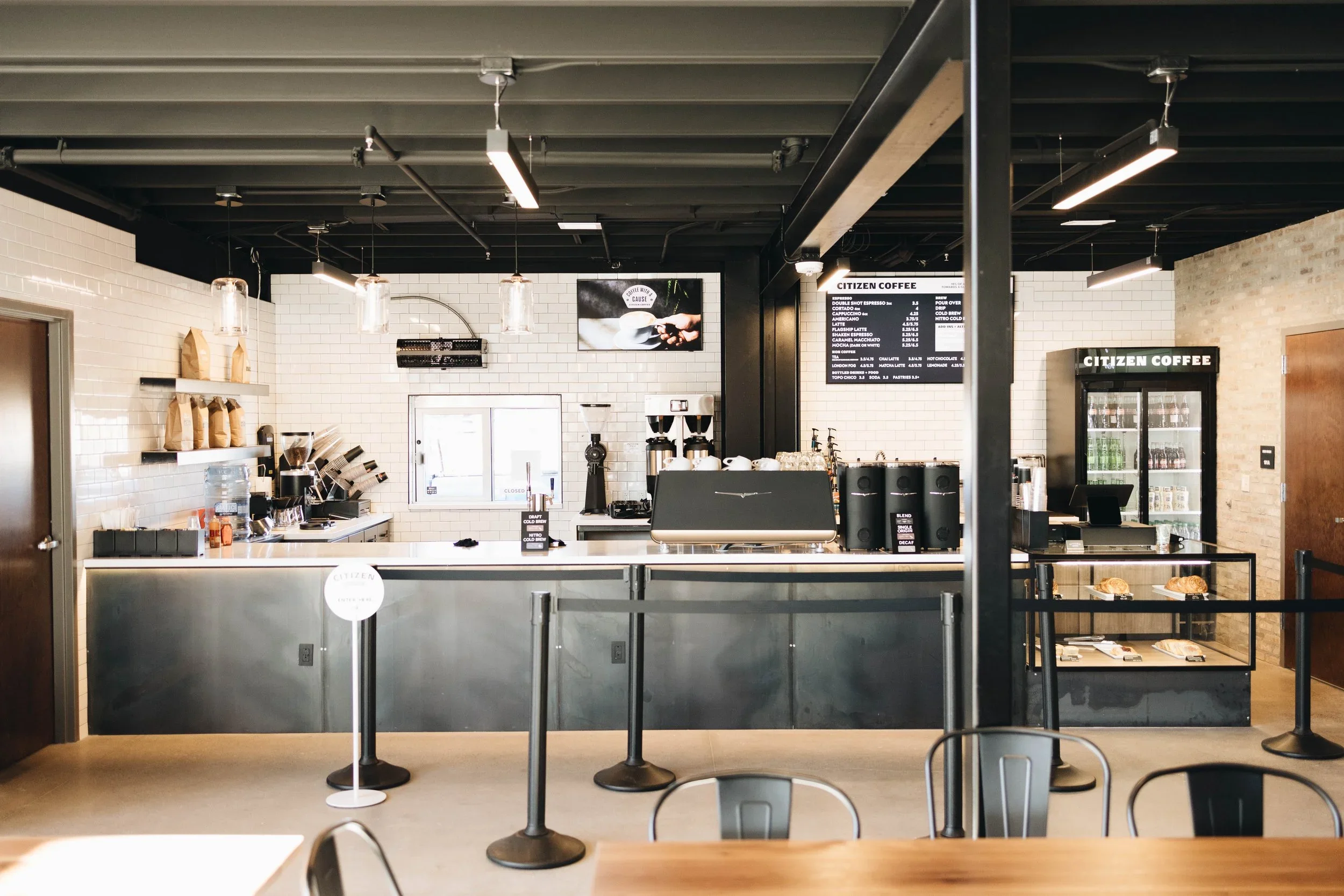 Interior of a modern coffee shop with white brick walls, black ceiling, menu board, coffee machines, refrigerated fridge, and display case with pastries.