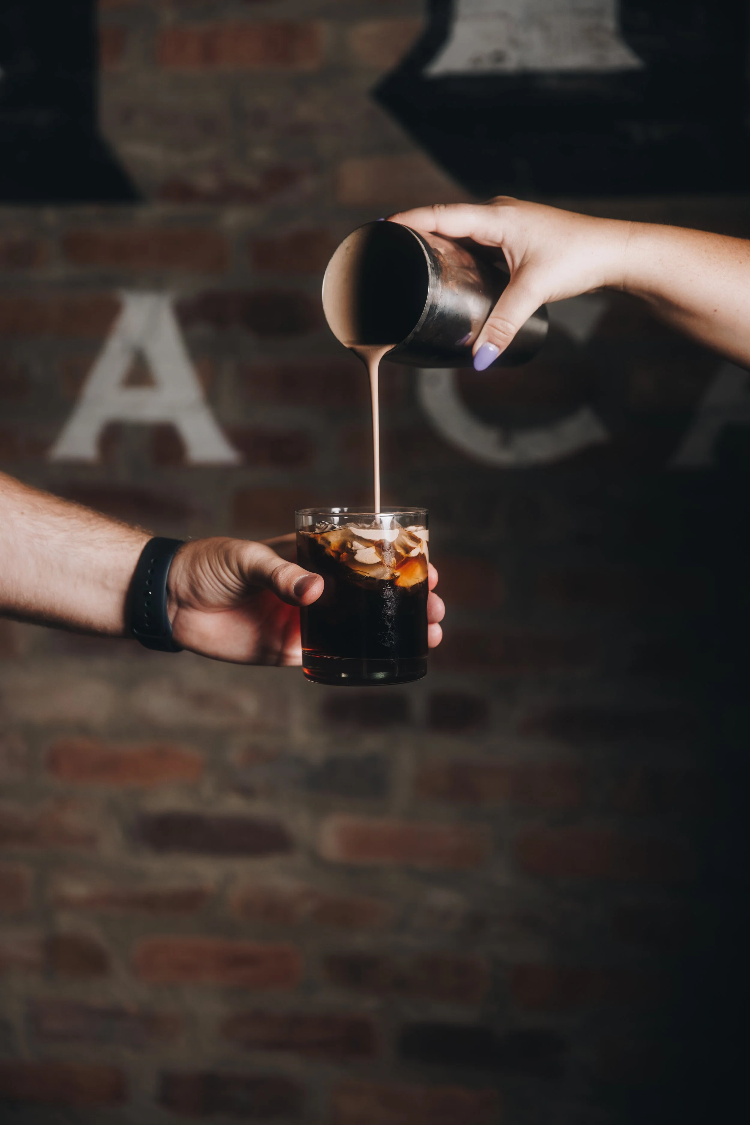 Person pouring milk into a glass of iced coffee with ice cubes against a brick wall background.