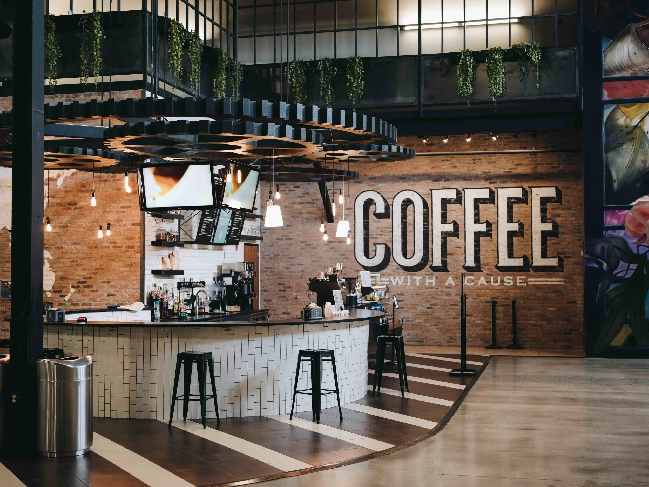 Interior of a modern coffee shop with a brick wall and large sign reading 'COFFEE' with the slogan 'with a cause'. There is a curved white tiled counter with bar stools and hanging lights, and a menu board behind the counter.