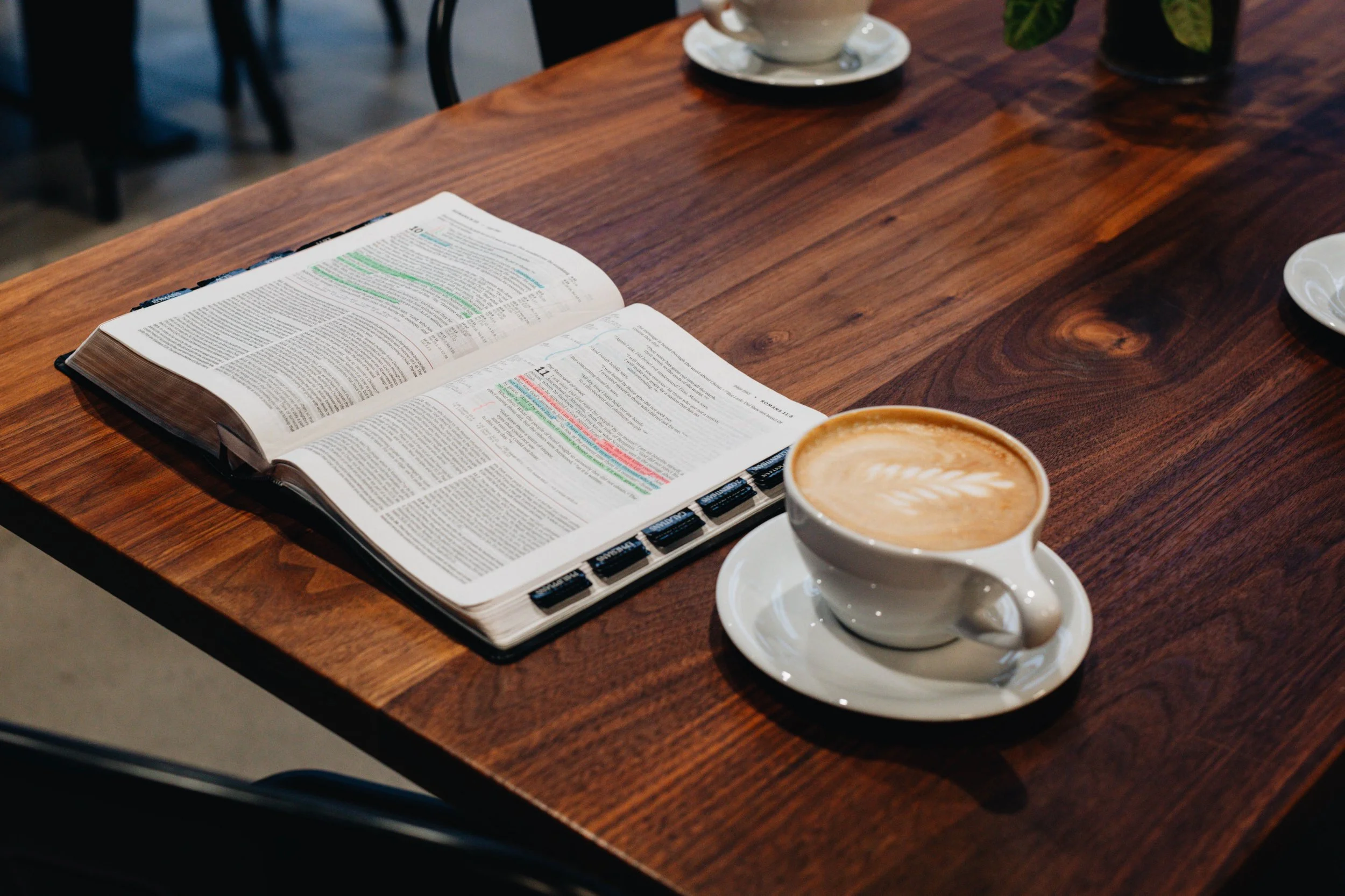An open book with highlighted text and tabs on a wooden table, next to a white cup of latte with a latte art leaf on top.