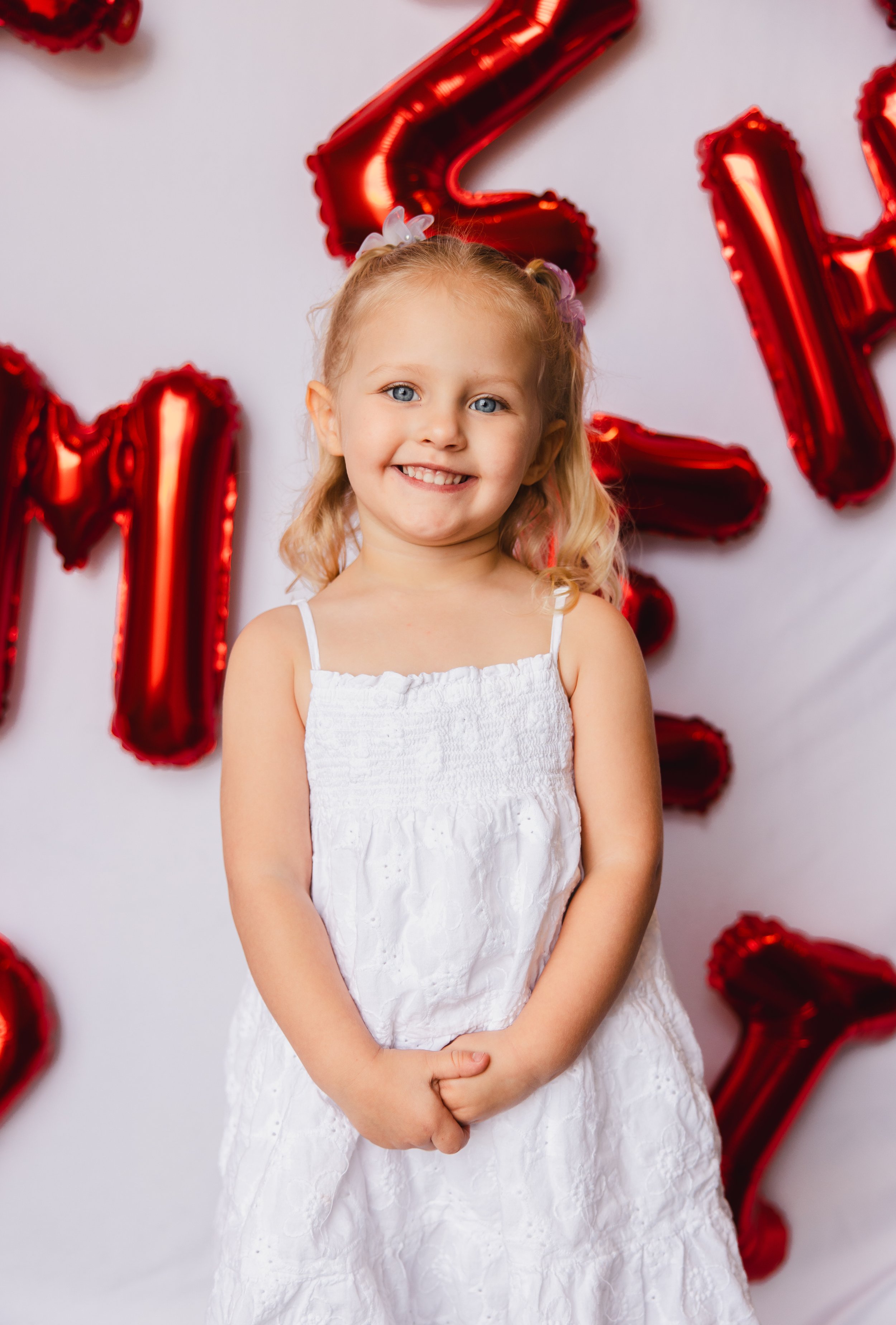 A young girl with blonde curly hair, wearing a white dress with spaghetti straps, smiling and standing in front of red foil balloons shaped like the number two and celebration letters.