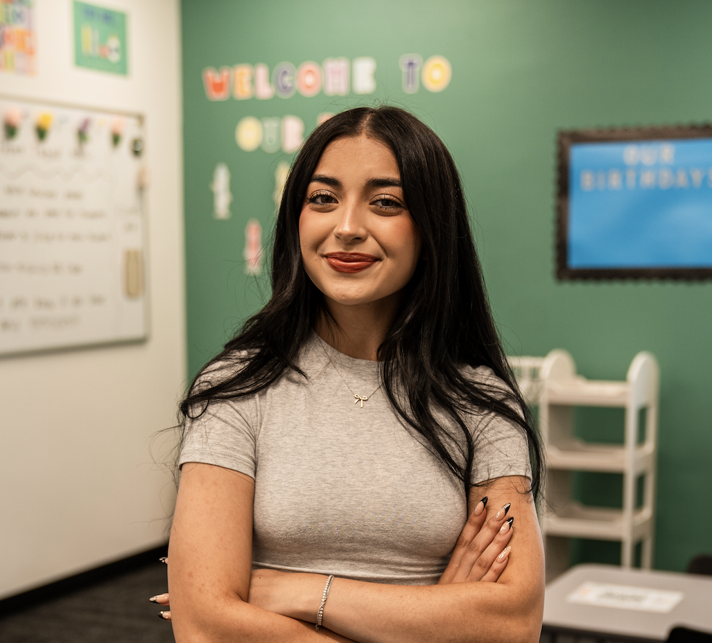 A young woman with long dark hair smiling inside a colorful classroom, standing with her arms crossed. The background includes a whiteboard and a bulletin board with the text 'WELCOME TO OUR' and other classroom decorations.