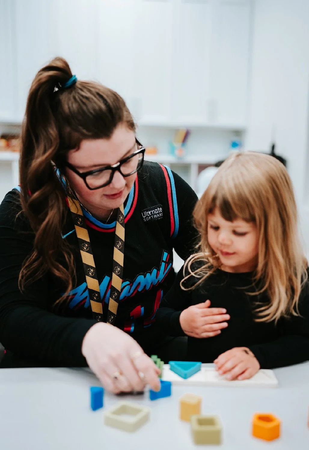 A woman with dark hair, glasses, and a lanyard teaching a young girl with blonde hair and a black shirt at a table with colorful geometric blocks.