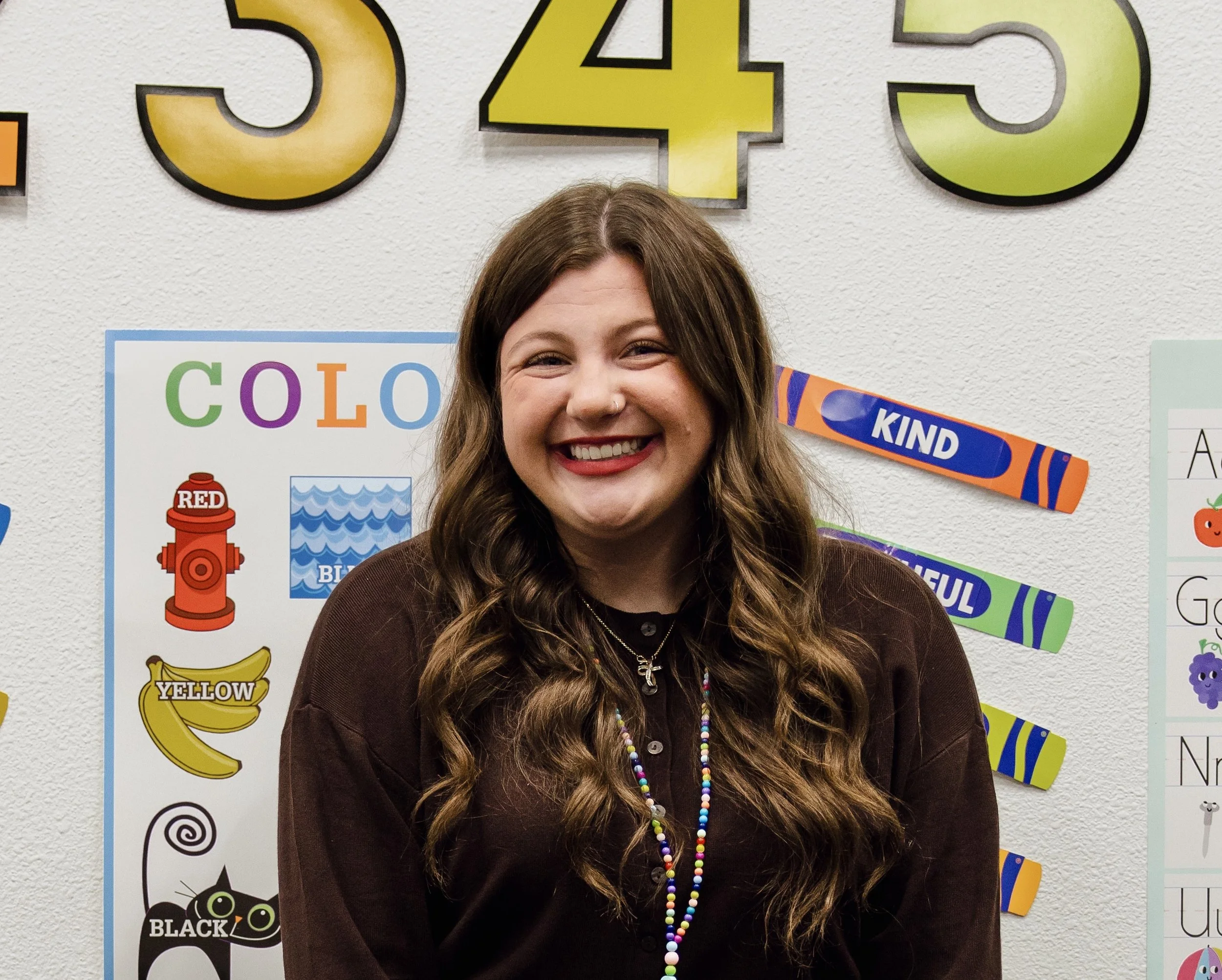 A woman with long wavy brown hair and red lipstick smiling, standing in front of educational posters and colorful letters on the wall.