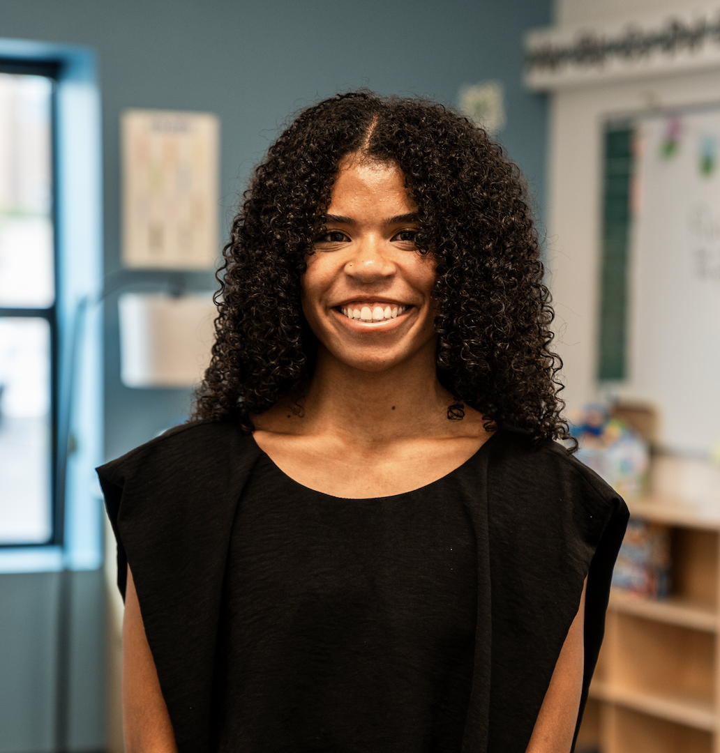 A smiling woman with curly black hair standing in a classroom or office with a bookshelf and window in the background.