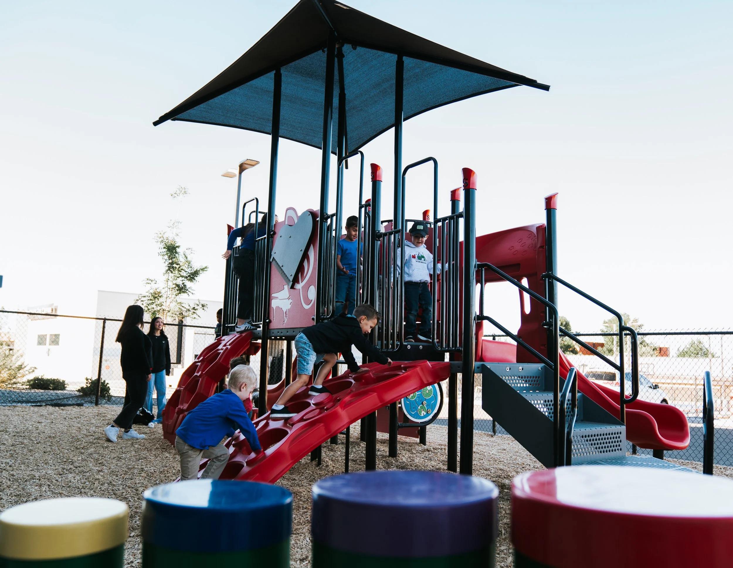 Children playing on a playground with a red slide and black canopy, surrounded by a chain-link fence on a sunny day.