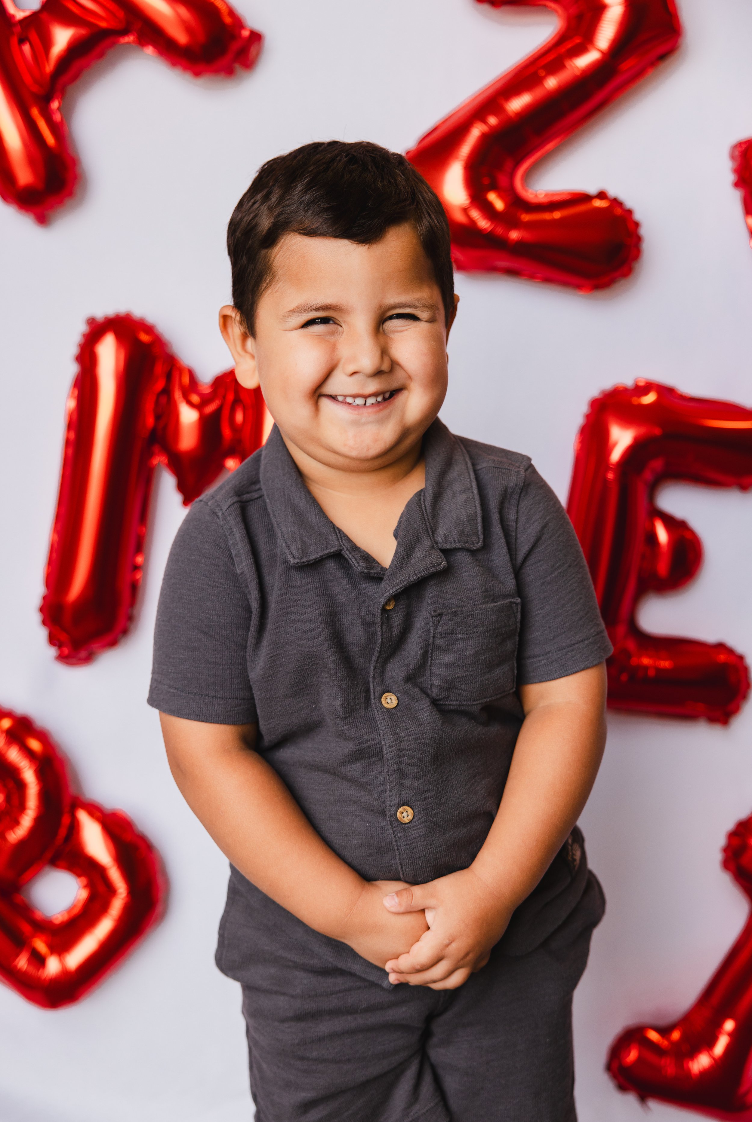 A young boy with dark hair, wearing a dark gray polo shirt, standing in front of a white background decorated with red foil balloons forming the number 2
