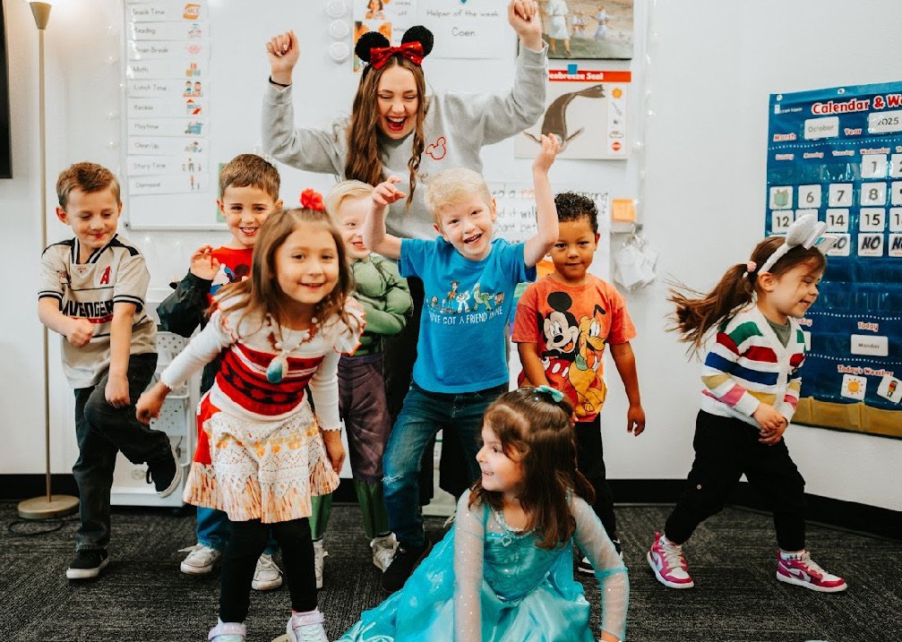 A group of young children and a female teacher are in a classroom. The children are smiling and posing energetically, some with raised fists, wearing colorful casual and festive clothes. The teacher is smiling and raising her arms in celebration. The classroom has educational posters, a calendar, and whiteboards in the background.