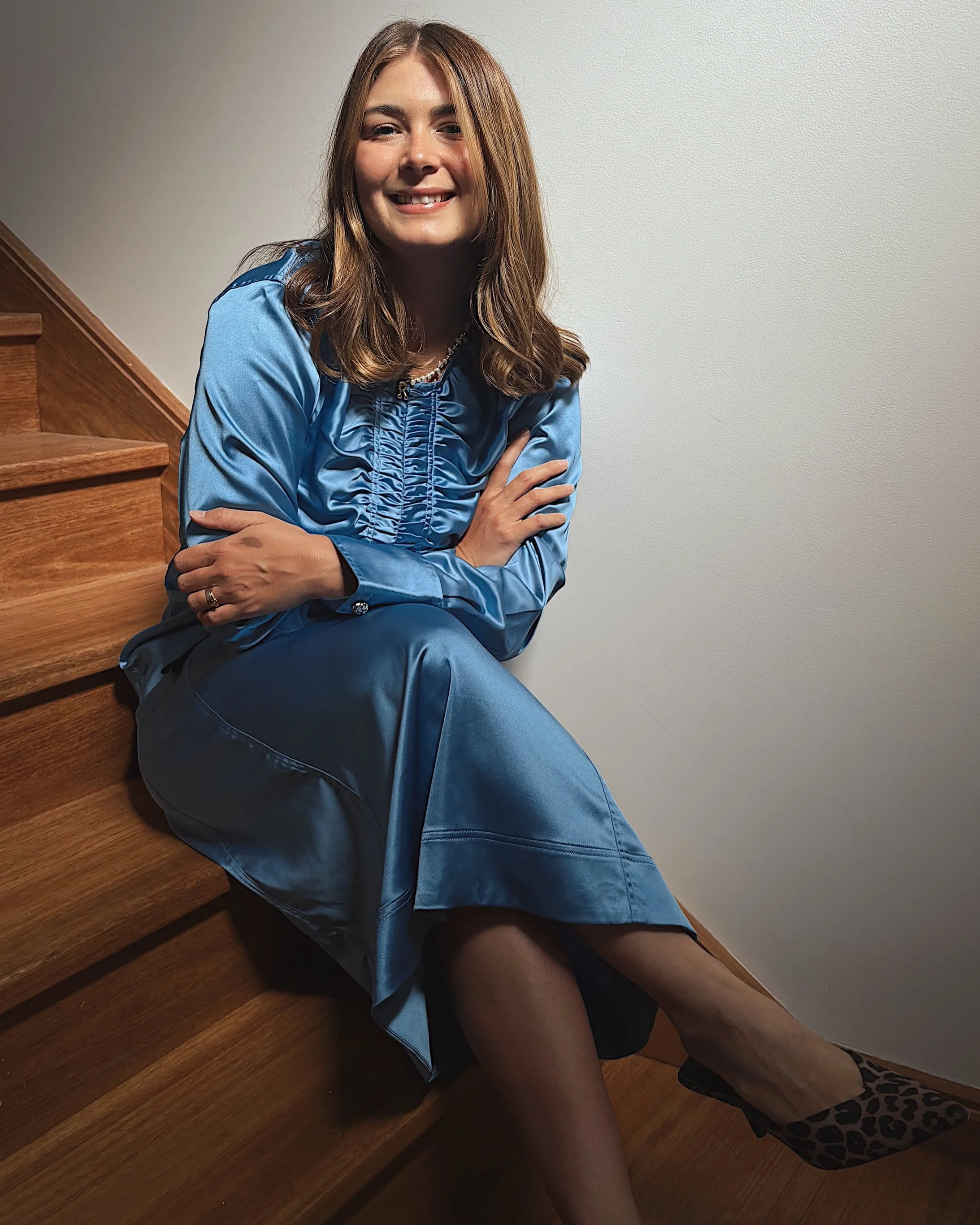 The business owner smiling while sitting on wooden stairs with her arms crossed. She is wearing a shiny blue dress, leopard print high heels, and a pearl necklace, against a plain wall background.