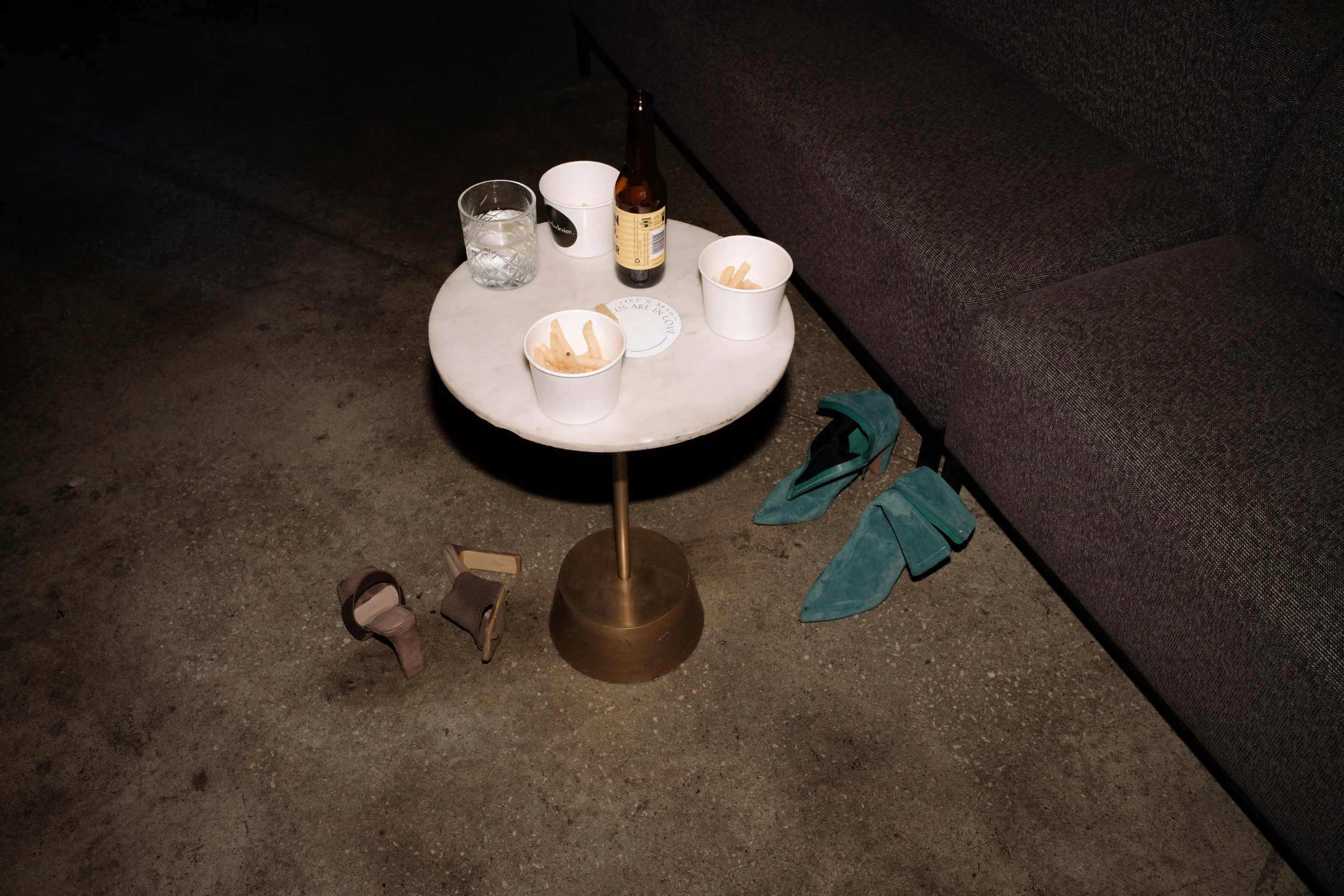 Empty marble-topped side table with snacks and drinks, surrounded by high heels on a concrete floor against a dark sofa.