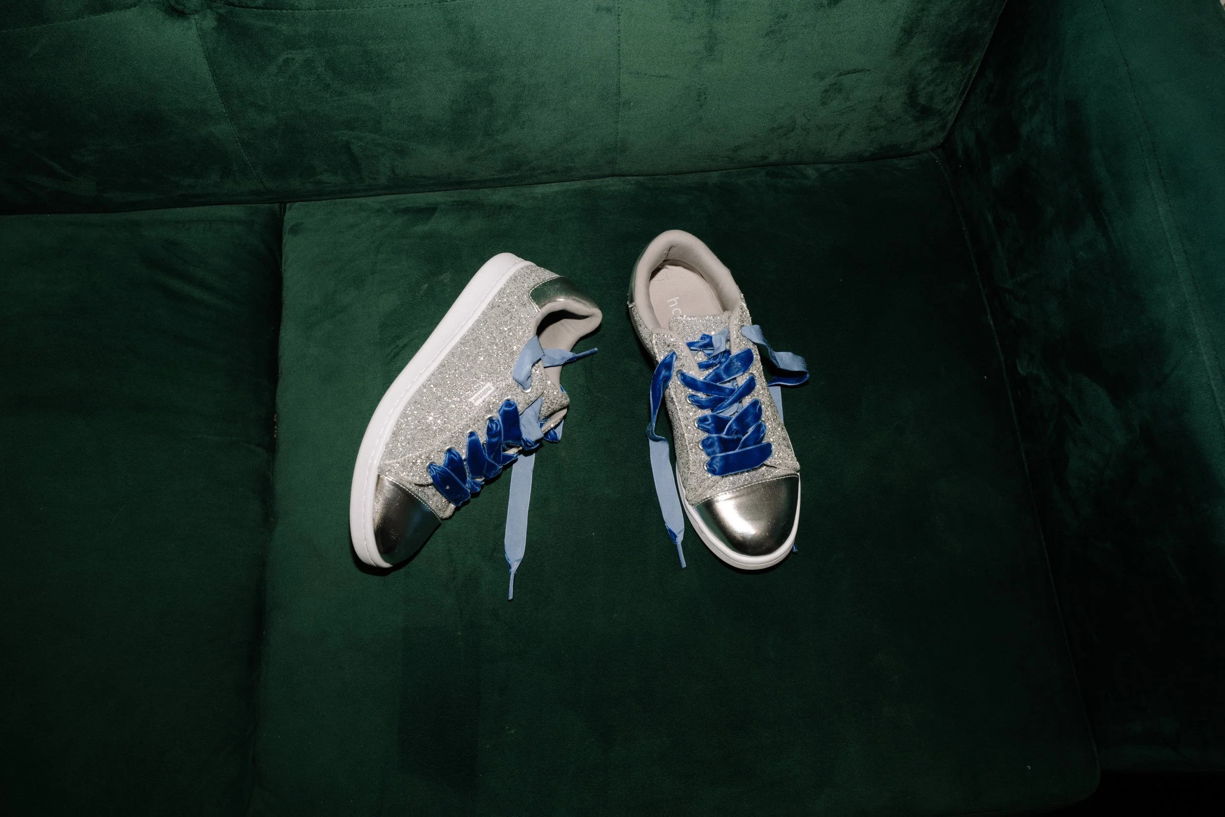 A pair of glittery silver sneakers with blue laces on a dark green sofa.