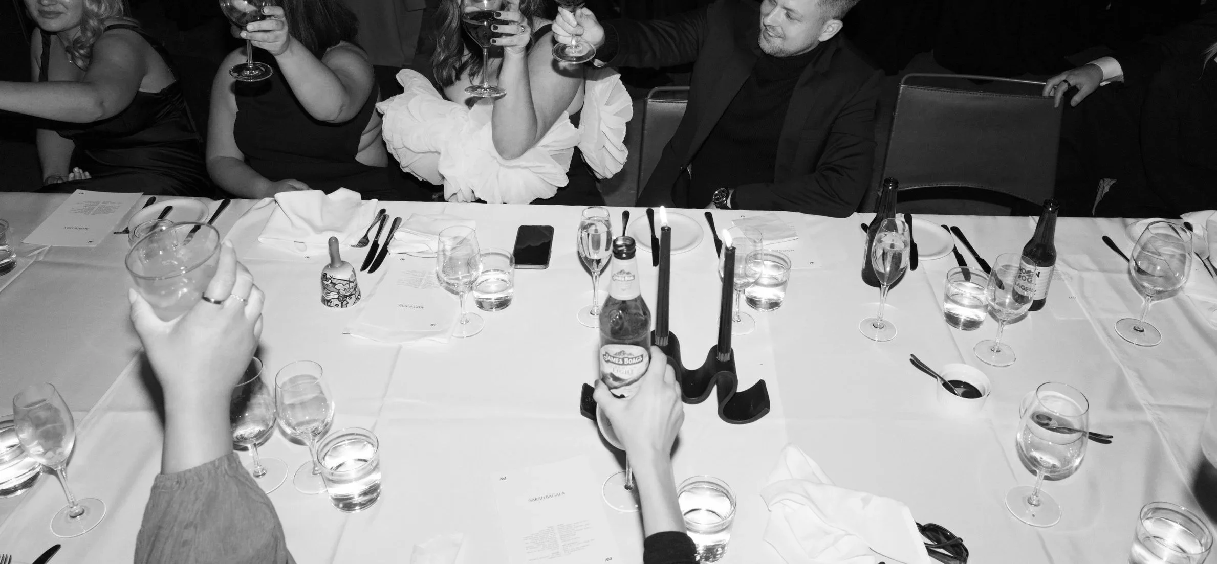People celebrating at a wedding party, raising glasses in a toast around a table with multiple glasses, bottles, and utensils.