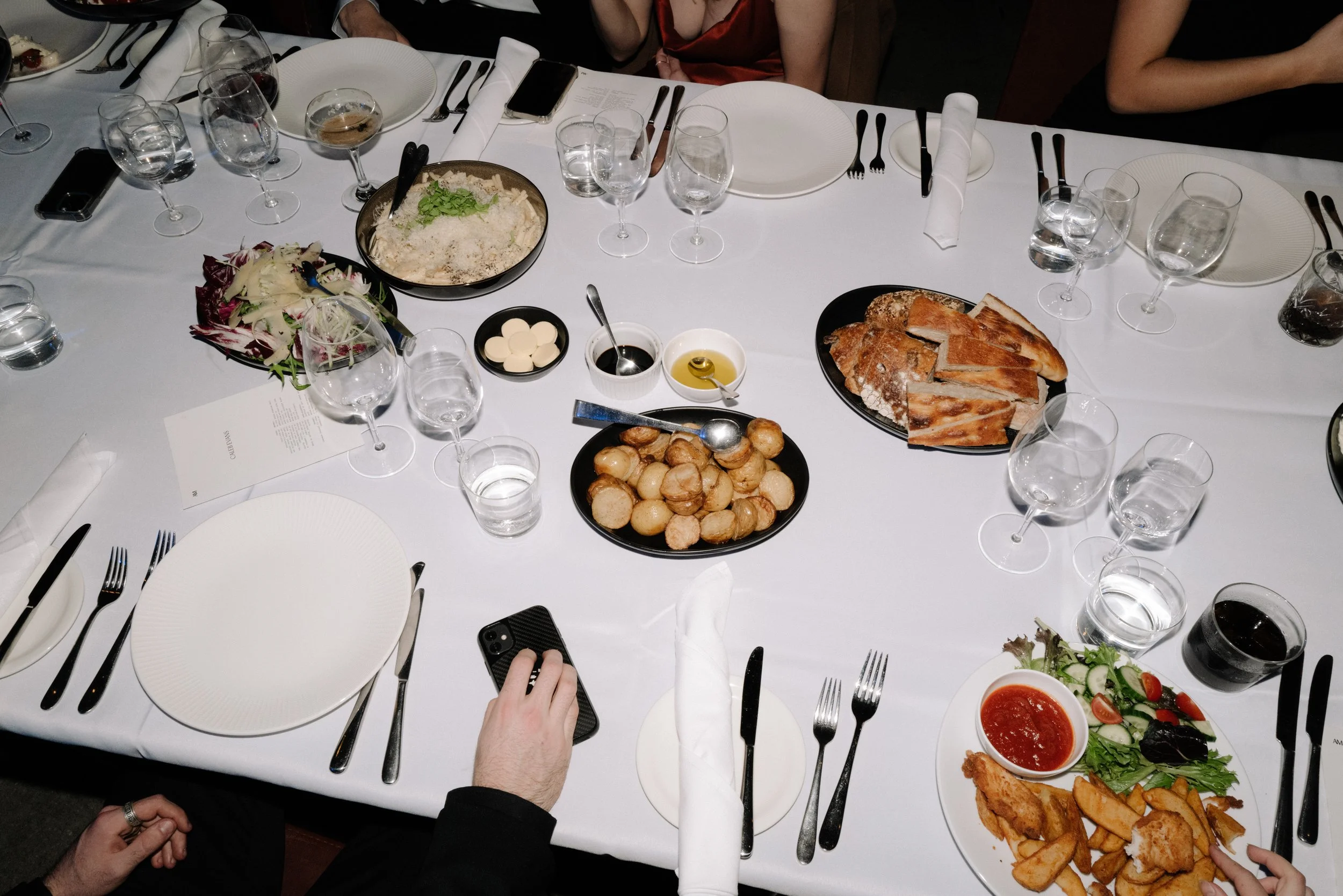 A formal dinner table set with plates, glasses, cutlery, and several dishes including bread, a salad, fried chicken with fries, and trays of other food. There are people seated around the table.