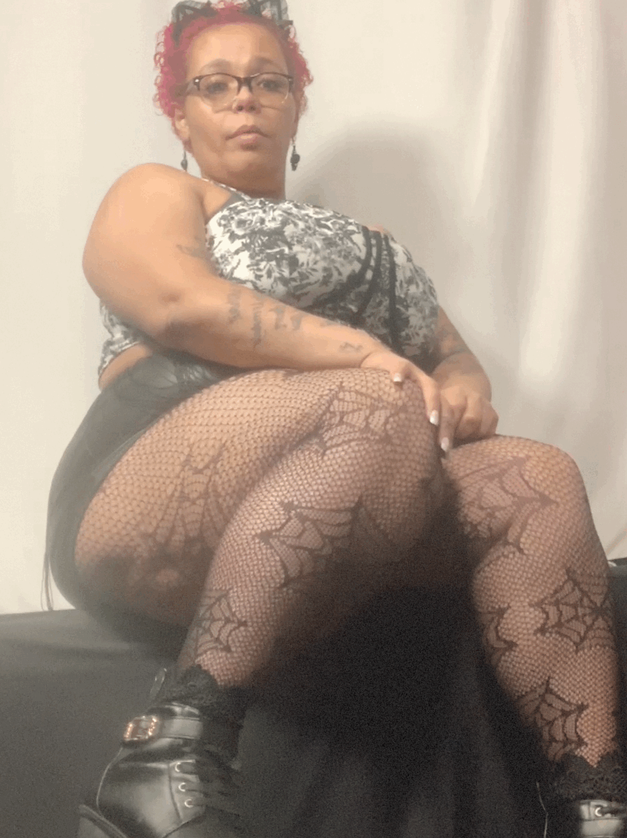 A woman with red hair and glasses sitting on a black surface, wearing fishnet tights with spiderweb patterns, a patterned top, and black boots, with a plain white wall and curtain in the background.